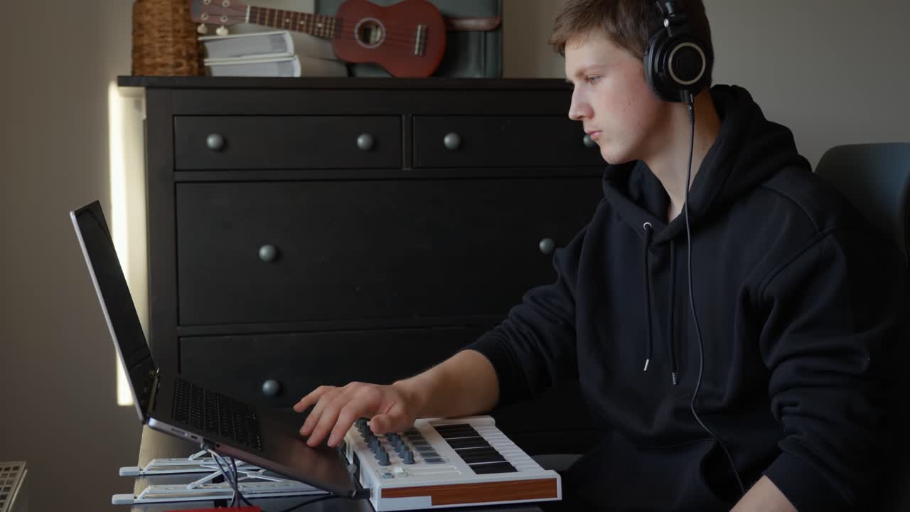 Young man wearing headphones producing music on a laptop and MIDI keyboard, working at home, focused on composing, arranging, and recording electronic audio tracks, static shot