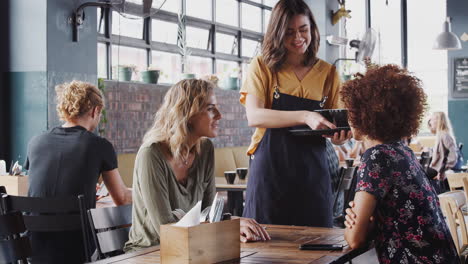 Two-Female-Friends-Sitting-At-Table-In-Coffee-Shop-Being-Served-By-Waitress