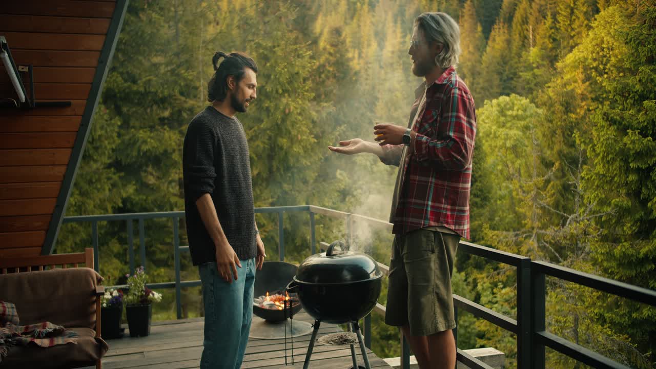 A couple of friends chat while cooking on the grill. A brunette guy in a green sweater lifts the lid of the grill and thick white smoke comes out. Picnic with a view of the mountains and coniferous forest