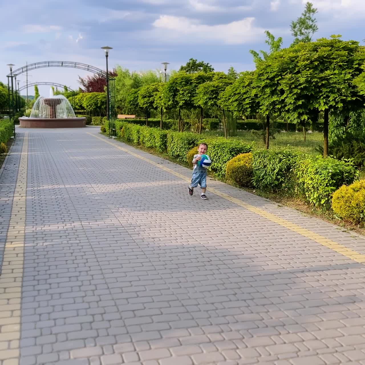Adorable little child holding a ball runs by the alley in the park. Kid throws the ball and follows it