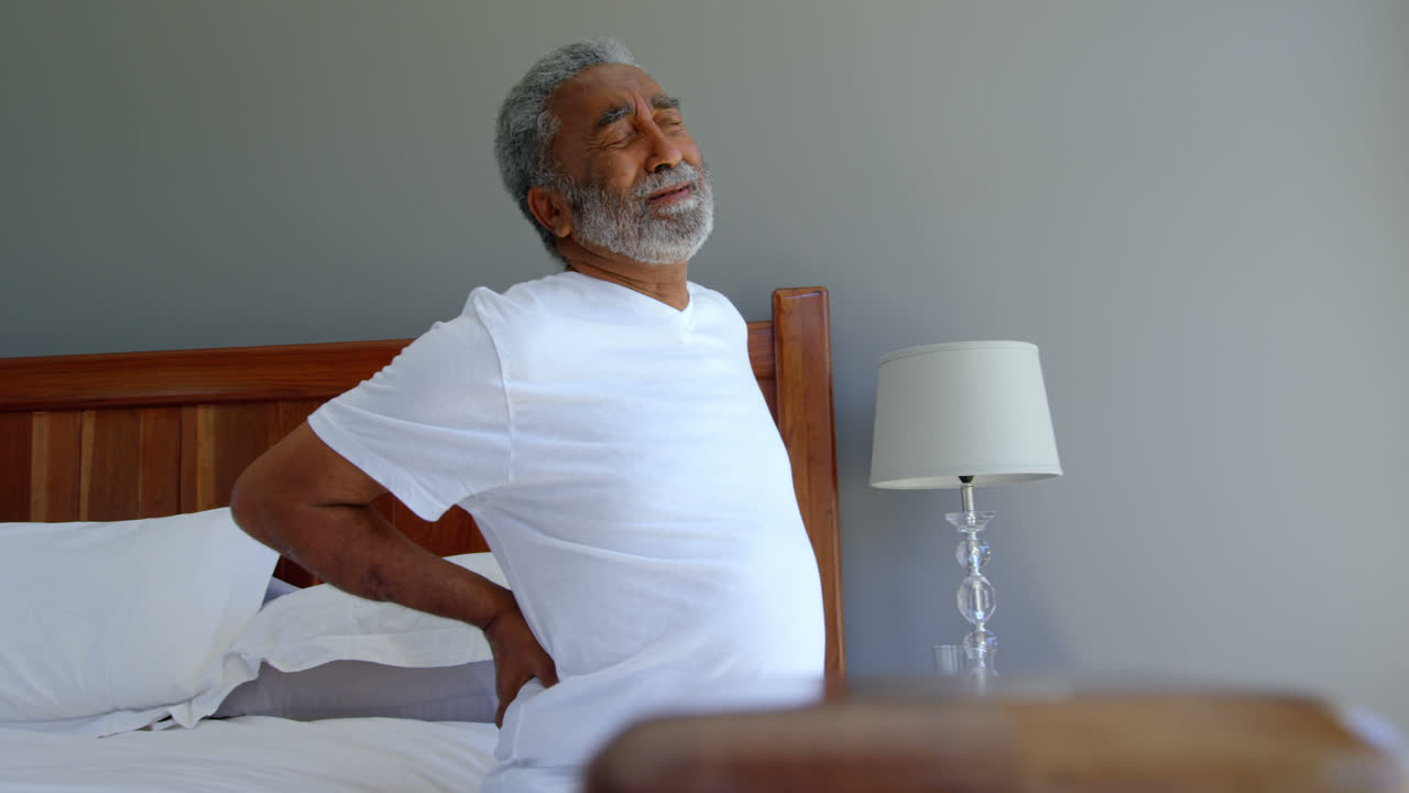 Front view of senior black man sitting on bed holding lower back in a comfortable home 4k