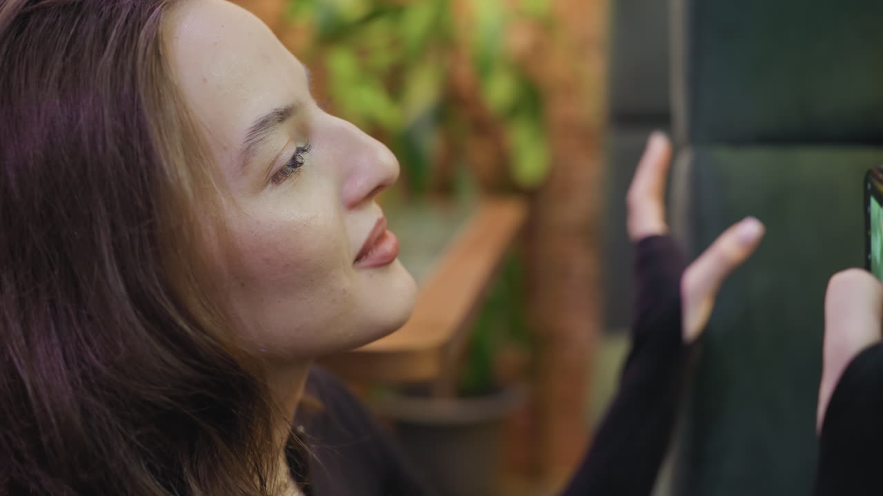 Close-up side view of woman with soft smile holding phone to take photo while leaning against cushioned chair, focused and calm with dim indoor light reflecting on her face and hands