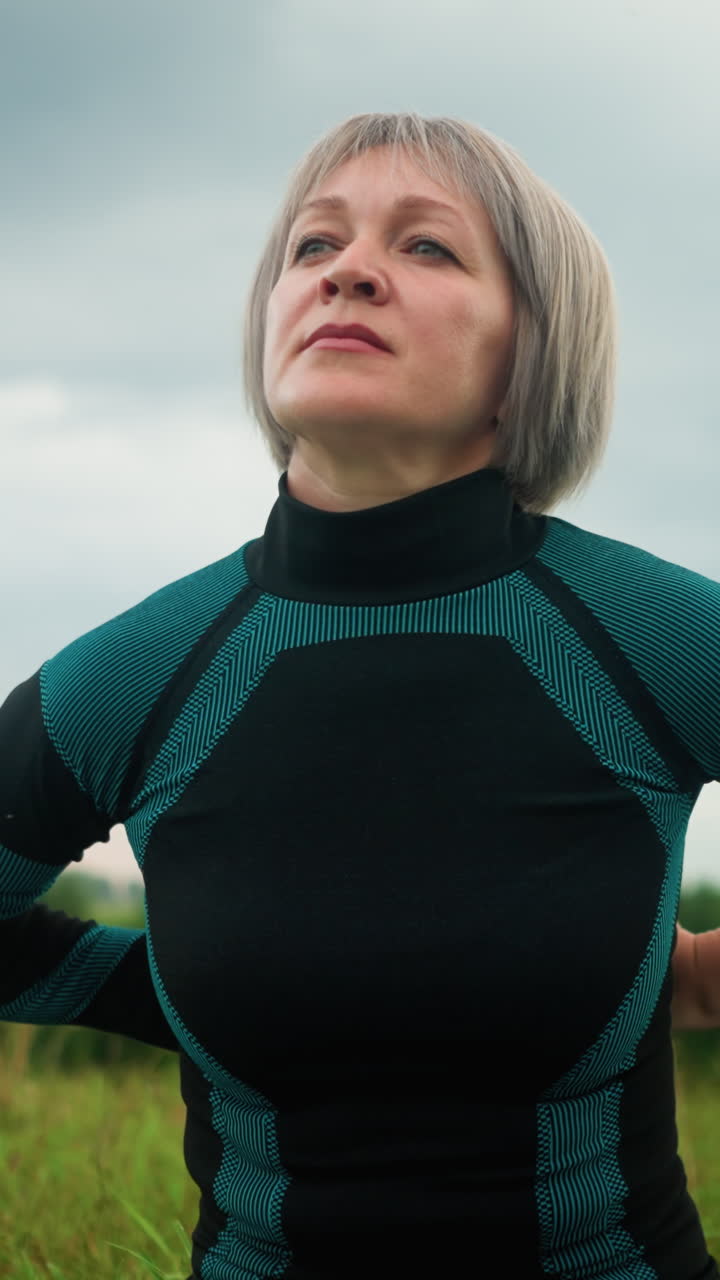 mujer de mediana edad sentada en sukhasana postura practicando yoga, mirando a la distancia con una expresión tranquila, en el fondo, los árboles borrosos están alineados bajo un cielo nublado