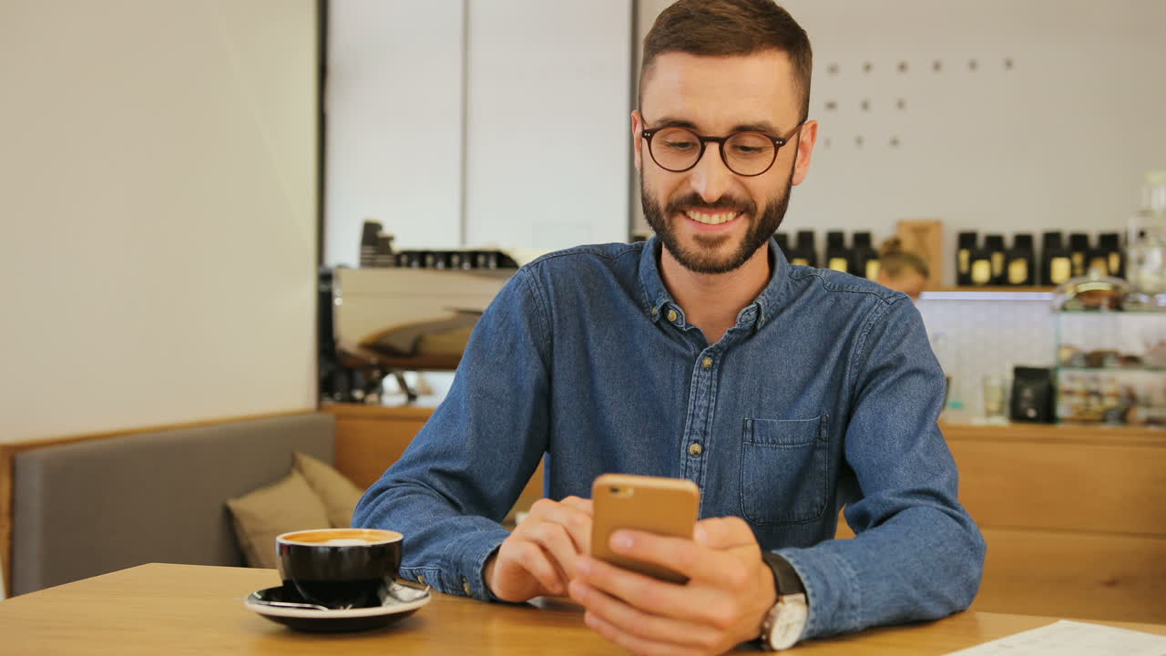 joven caucásico con anteojos usando un smartphone para ver un video divertido mientras se sienta en una mesa en un café