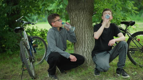 two young siblings squatting under a tree, drinking from water bottles, while resting after a bicycle ride, both bicycles are parked nearby, surrounded by lush greenery in a peaceful forest