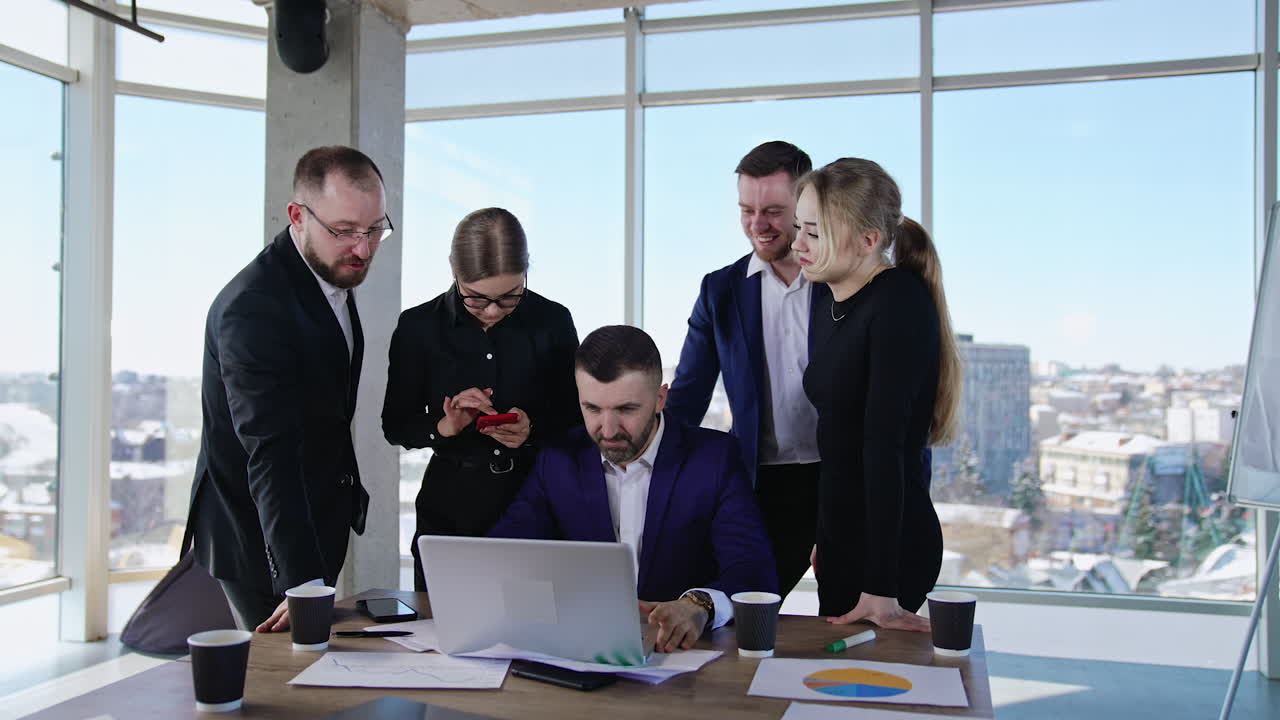 Male leader sitting in front of laptop surrounded by his teammates. Businesspeople communicating at work. City view in the windows of office.