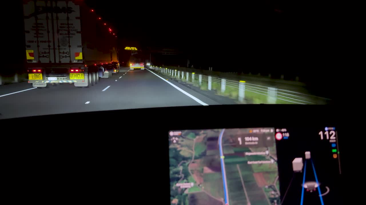 Car follows truck on illuminated highway at night, dashboard navigation screen visible, steady camera perspective