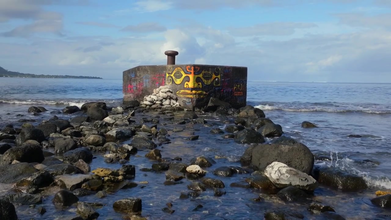 Fransız Polinezyası, Tahiti, Teahupoo'ya giden Taiarapu yolu üzerinde deniz kenarında sokak sanatı. Güney Pasifik Okyanusu'nda drone çekimi. Çimento blok üzerine Polinezya yüzü grafiti resmi, alçak gelgit kayaları, çarpan dalgalar, güneşli bir gün ve ileri dolly zoom tekniği.
