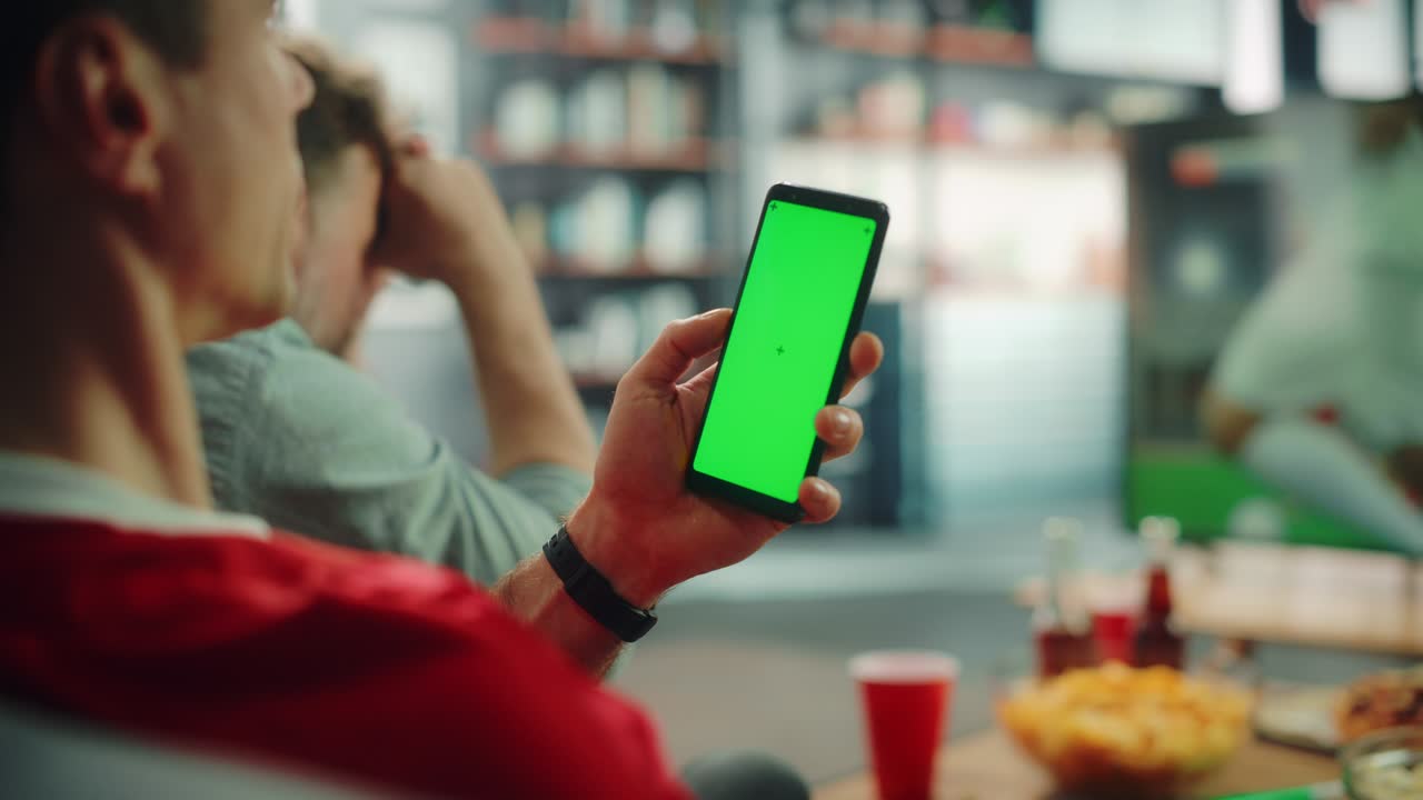 en casa fan de fútbol sostiene pantalla verde chroma clave teléfono inteligente, con amigos ve el juego de fútbol en la televisión, animar al equipo deportivo favorito para ganar el campeonato. juego de club de fútbol. sobre el hombro