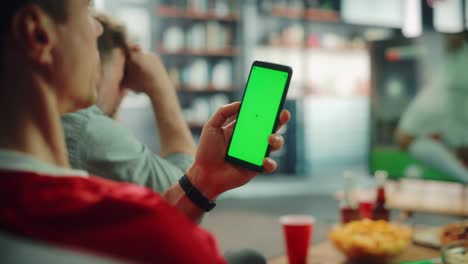 en casa fan de fútbol sostiene pantalla verde chroma clave teléfono inteligente, con amigos ve el juego de fútbol en la televisión, animar al equipo deportivo favorito para ganar el campeonato. juego de club de fútbol. sobre el hombro