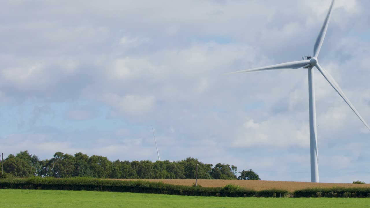 A single wind turbine spins steadily in a green field near Edinburgh, Scotland, under partly cloudy daylight. Static wide shot with natural lighting