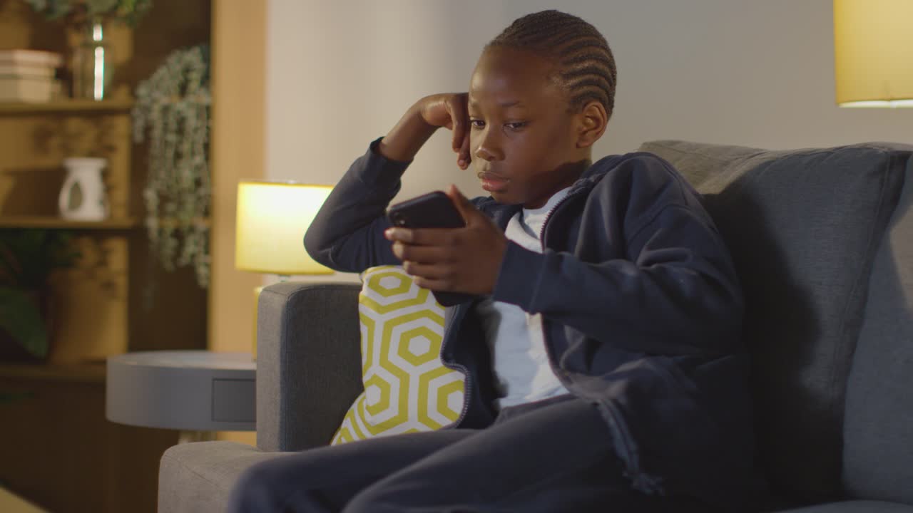 Garçon à la maison assis sur un canapé le soir, regardant la télévision et consultant son téléphone portable.