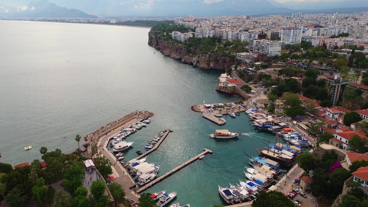 Ripresa aerea con drone in tracking sopra lo storico porto romano del quartiere di Kaleici, che rivela barche ormeggiate lungo il molo di pietra curvo sotto ripide mura cittadine ad Antalya, Turchia