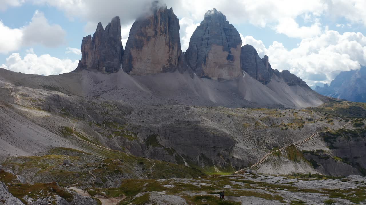 Vue aérienne de randonneurs marchant le long d'un sentier avec les Tre Cime di Lavaredo et des nuages sur les sommets au loin.