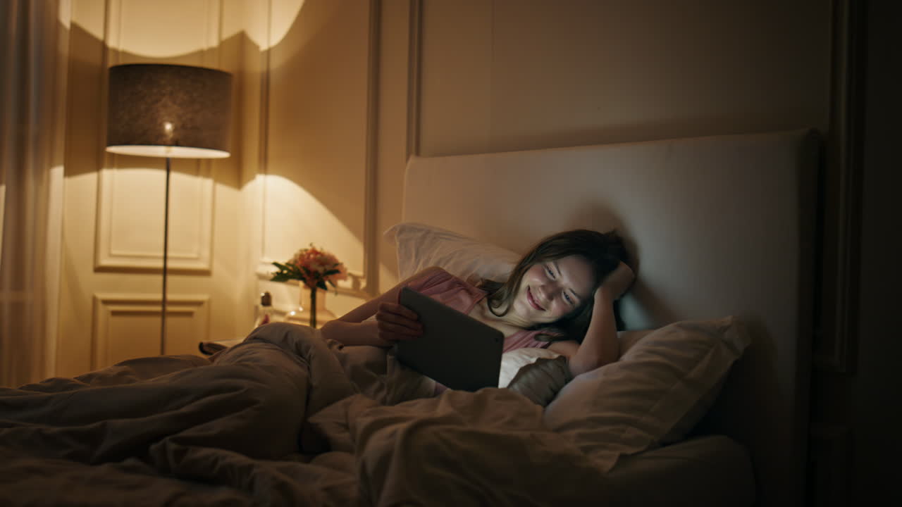 Relaxed girl looking tablet in cozy bed at night. Smiling happy woman watching