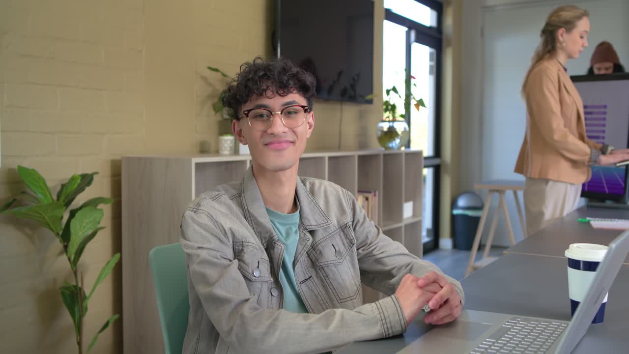 Impiegato che digita al computer portatile alla scrivania in ufficio, nota la telecamera e ride con una tazza di caffè in mano