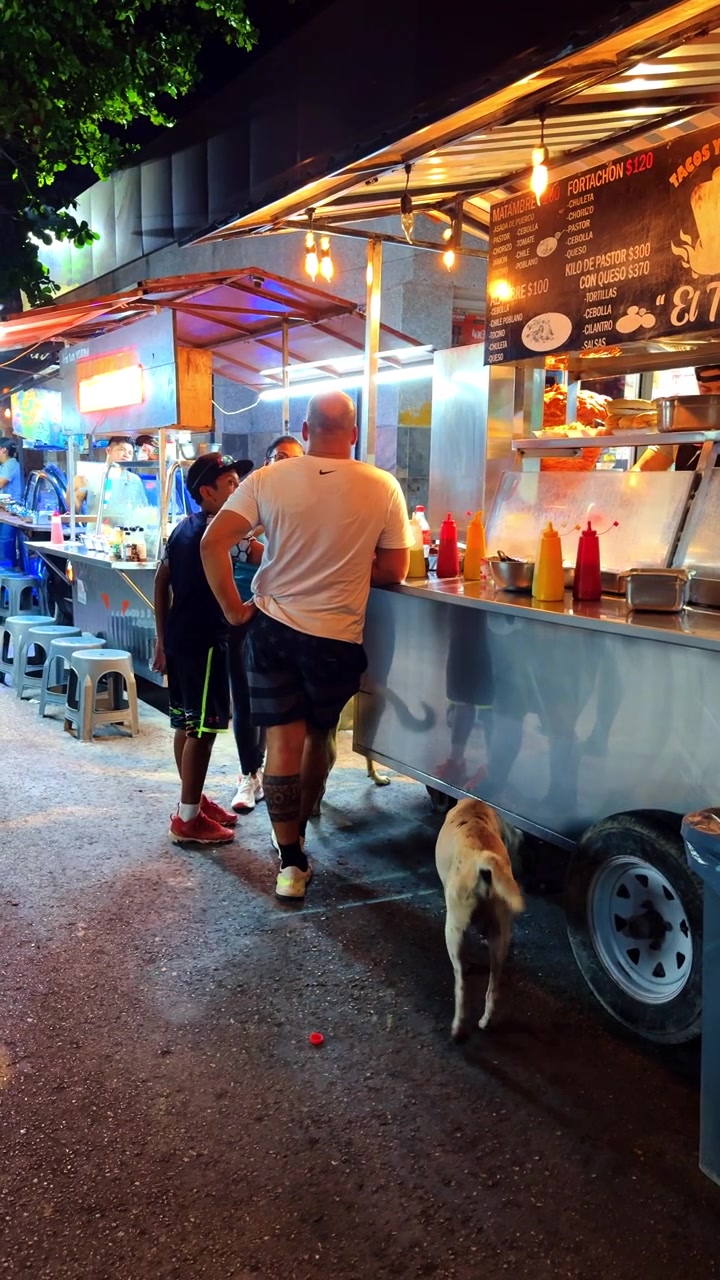Tulum, Messico, 1 agosto 2025: Persone in attesa a una bancarella di street food notturna in Messico. Scena notturna di clienti in piedi accanto a una bancarella di taco in acciaio inossidabile con tabellone del menu e bottiglie di salsa colorate.
