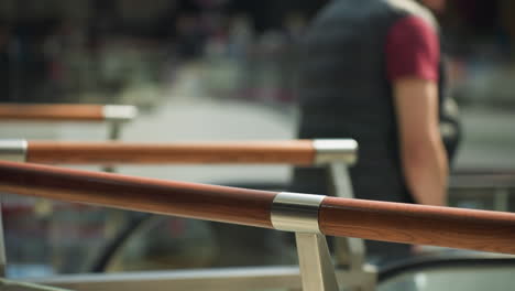 a bustling mall scene captured from a first-person perspective with a handheld camera, featuring a blurred background. the focus is on sleek wooden handrails with a person holding onto them