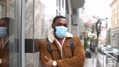 black man with medical mask and shoulder backpack stands outdoor near the office center