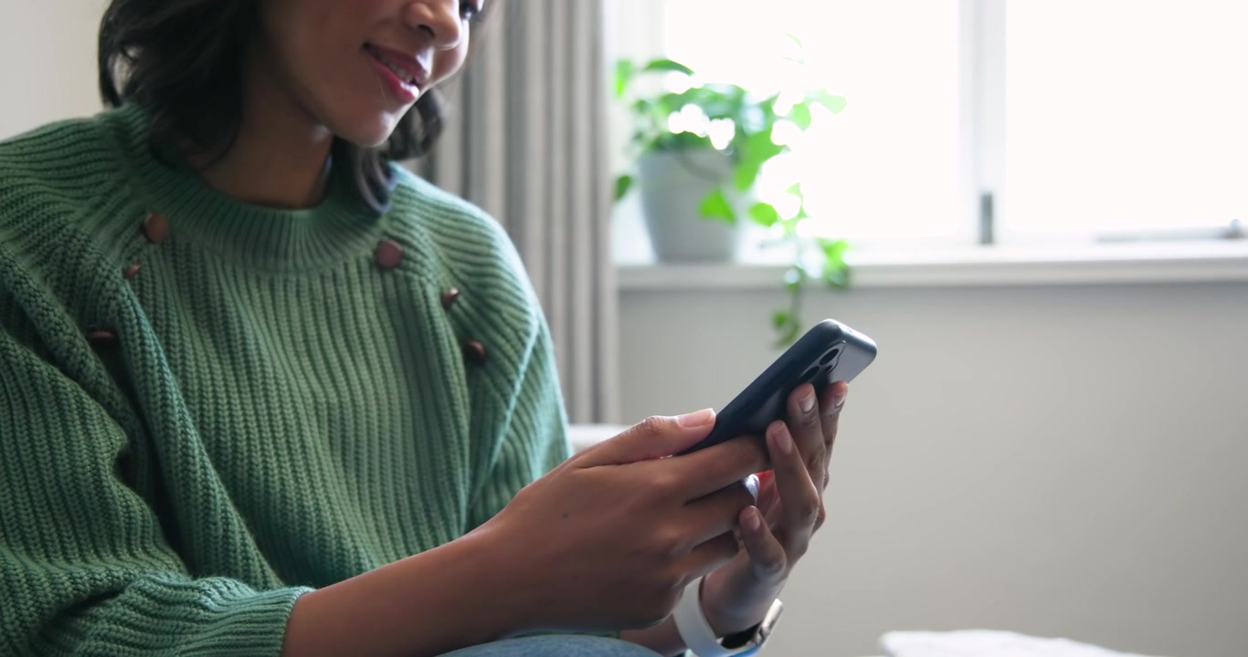 Afroameričanka drží telefon, ťuká palci, pohlíží na rostlinu u okna a nosí chytré hodinky.