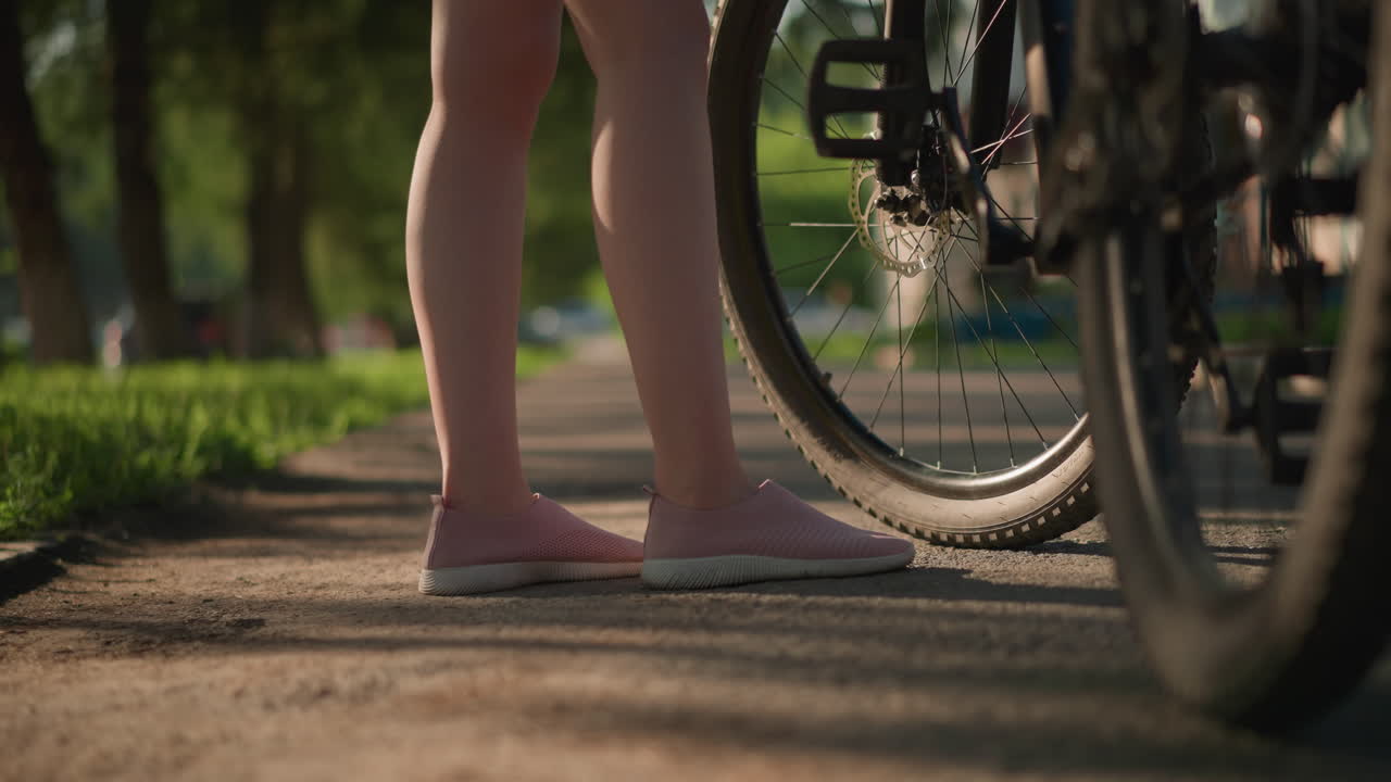 Close-up of individual legs in pink sneakers kicking a bicycle tire to check air pressure, lush greenery and trees create a beautiful backdrop while a car passes by in the distance