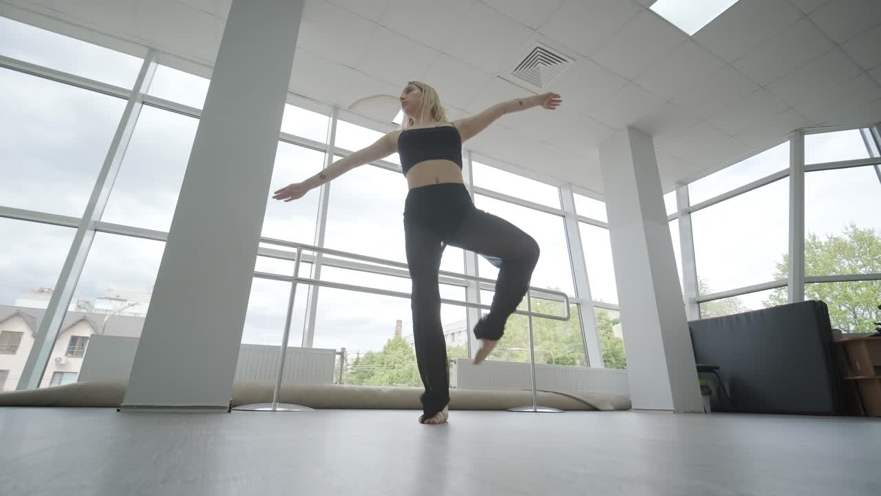 Fit girl in black clothes performing contemporary. Female dancer shows flowing motions, body and hands waves at white room. Modern ballet dance choreography. Young woman is moving smoothly
