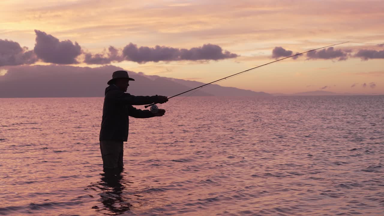 Gerak lambat digunakan untuk menunjukkan bidikan penuh pemancing lalat dalam siluet saat matahari terbenam di Molokai, Hawaii.
