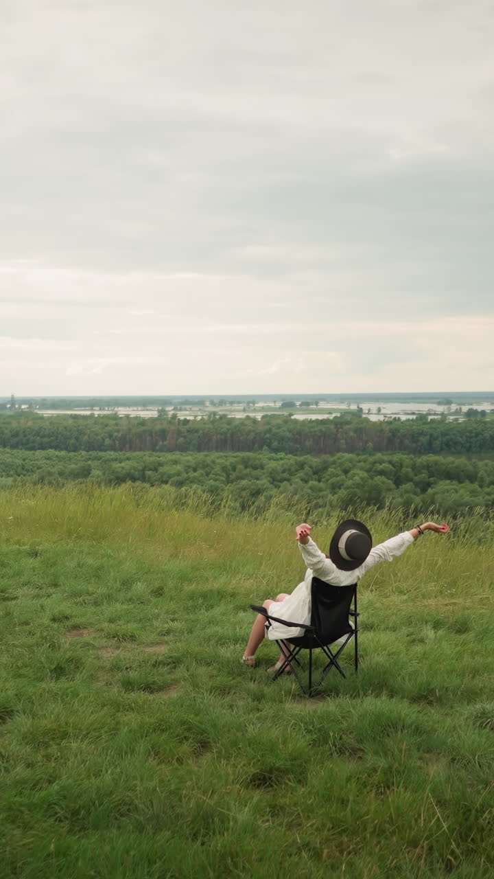 A serene scene capturing a woman in a black hat, sitting comfortably on a chair with her hands touching her ears while an artist paints her in a lush grassy field by a tranquil lake