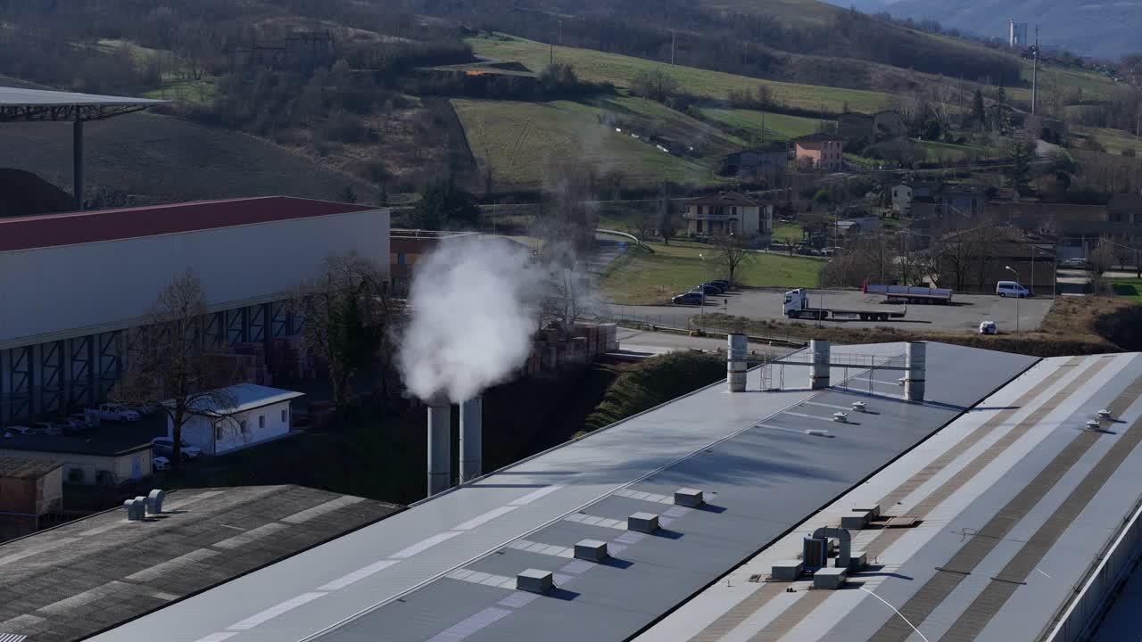 Impianto industriale con tetti in metallo e camini che emettono vapore bianco nell'aria in un'area di produzione industriale, ripresa aerea teleobiettivo con drone.