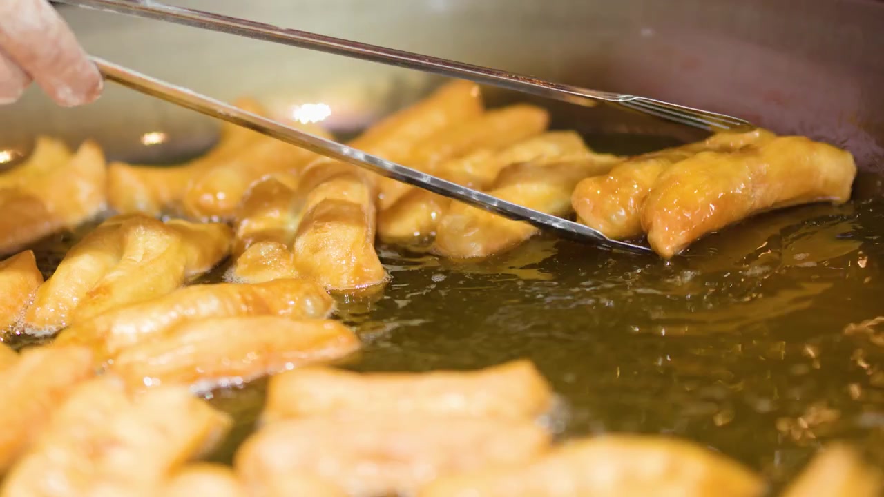 Gros plan de bâtonnets de pâte dorés frits dans de l'huile bouillonnante à l'aide de pinces métalliques