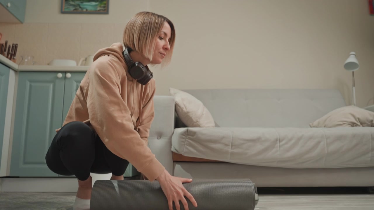 White woman unrolling yoga mat headphones around neck closeup on textured foam and floor, hands smoothing surface, minimalist apartment kitchen, focused warmup ritual before workout