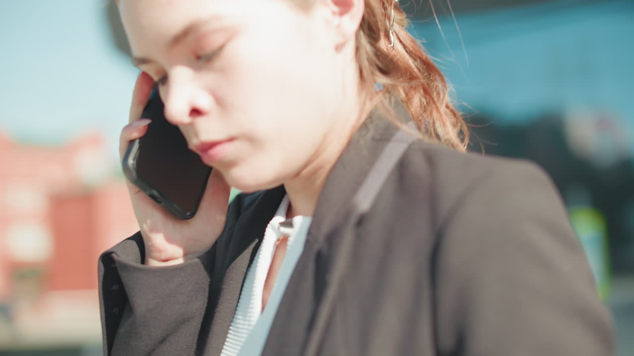 Angry woman on phone drops device in frustration while continuing call, exiting building dressed in formal attire, standing outside glass door with urban background