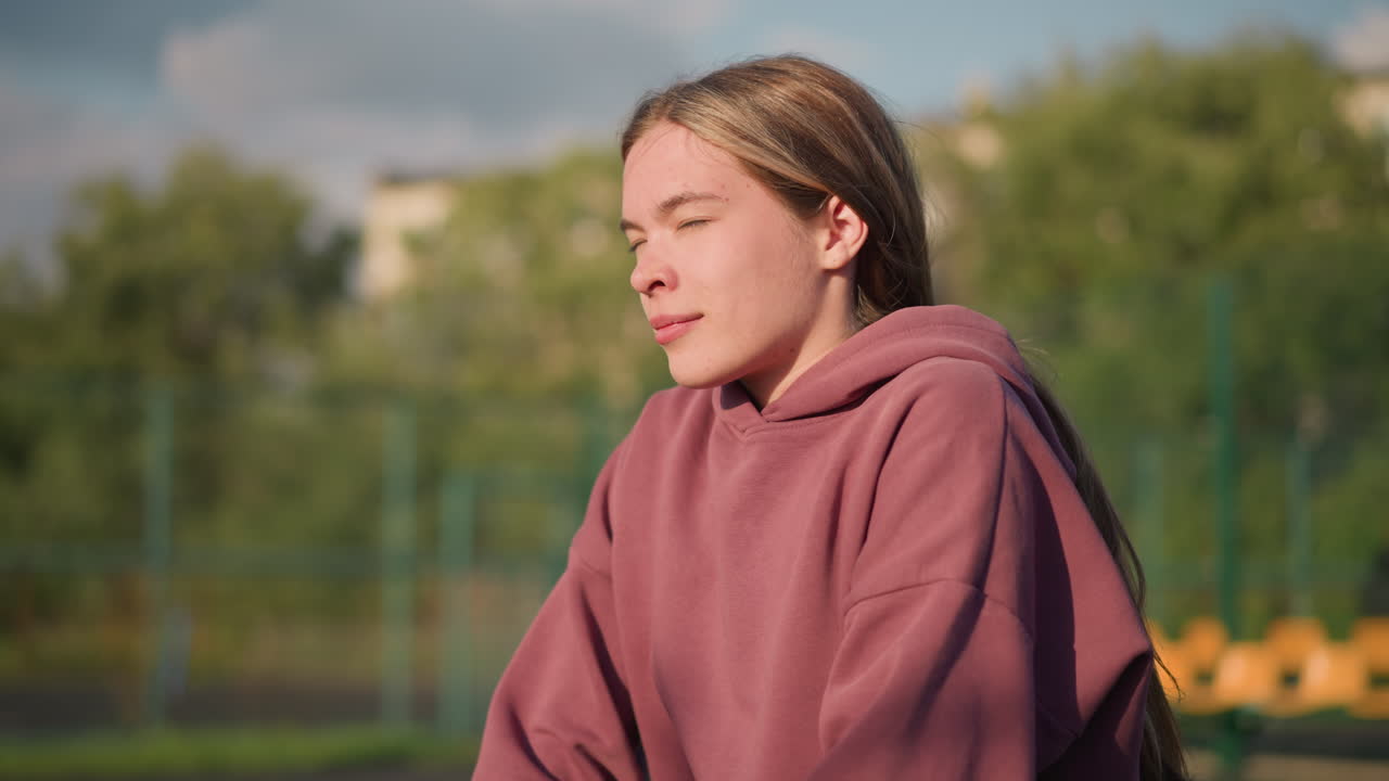 피트니스 운동에 참여하는 운동선수, 앞으로 무을 고 머리카락을 뒤로 고, 야외 스포츠 환경에서 나무와 불분명한 인물이 있는 흐릿한 배경으로 아침 드릴을 즐기고 있습니다.