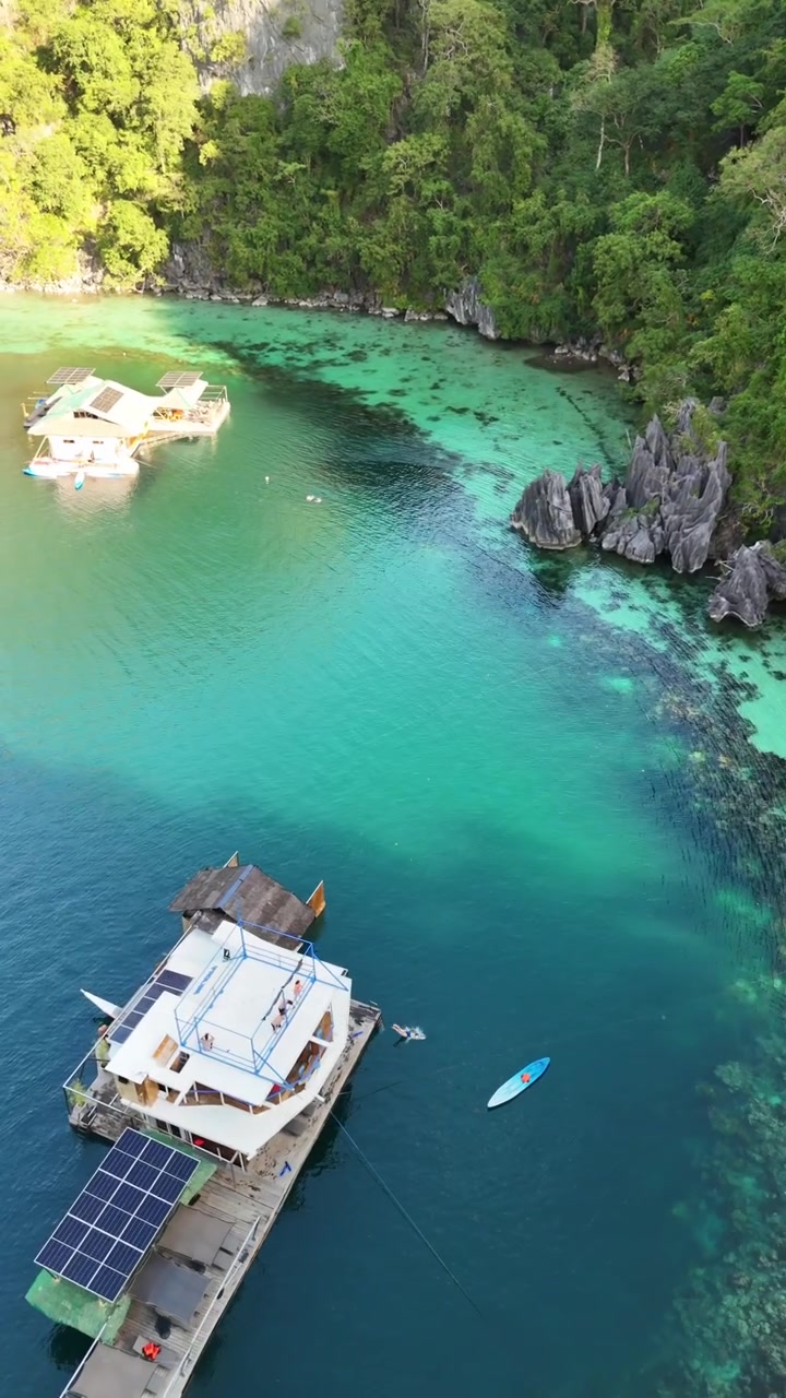 Pemandangan udara sudut tinggi yang menakjubkan dari rumah perahu apung bertenaga surya yang berlabuh di air laguna pirus jernih, dikelilingi tebing hutan lebat dan papan dayung di Palawan, Filipina
