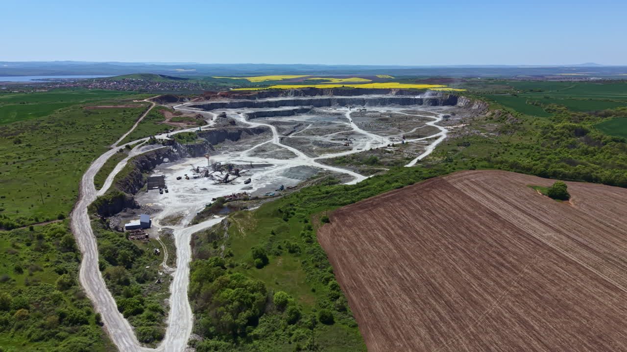 Grande sito minerario visto dall'alto con macchinari in funzione circondato da campi sotto un cielo sereno di giorno.