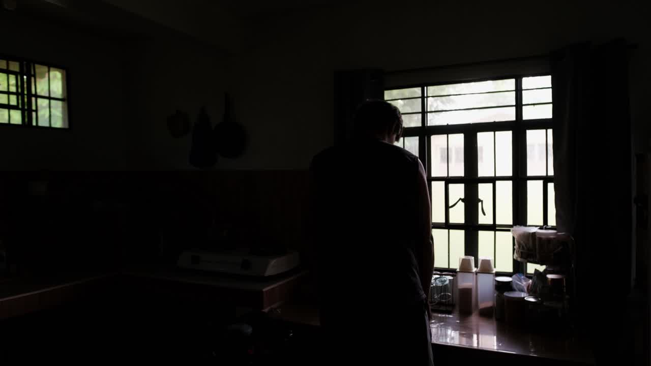 Rear static shot of young man walking into apartment kitchen and opening the window curtains, revealing morning sunlight