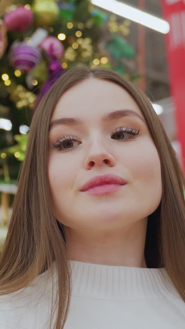 Stylish lady takes a picture with her phone in front of vibrant Christmas tree and decor at a well-lit holiday store, she adjusts her hair, with shoppers blurred in the background