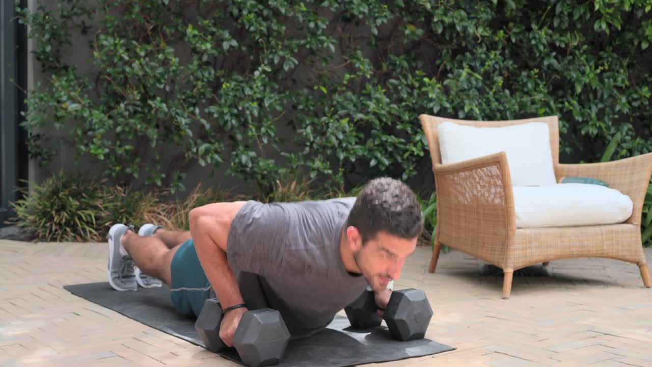 Man in sportswear doing push-ups starting lowered on mat on brick patio with dumbbells for strength