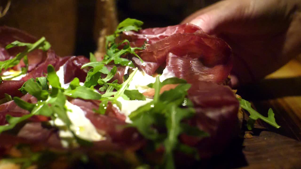 Hand picking up a piece of pizza with bresaola, burrata, and arugula