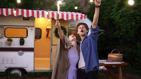 Young-Couple-Embracing-Standing-In-Front-Trailer-They-Living-In-With-Sparklers-Celebrating,-Dancing-And-Smiling