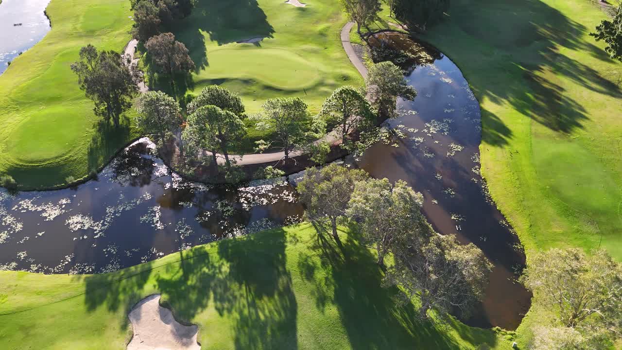 Drone footage captures a lush golf course with water hazards and trees, bathed in golden sunset light