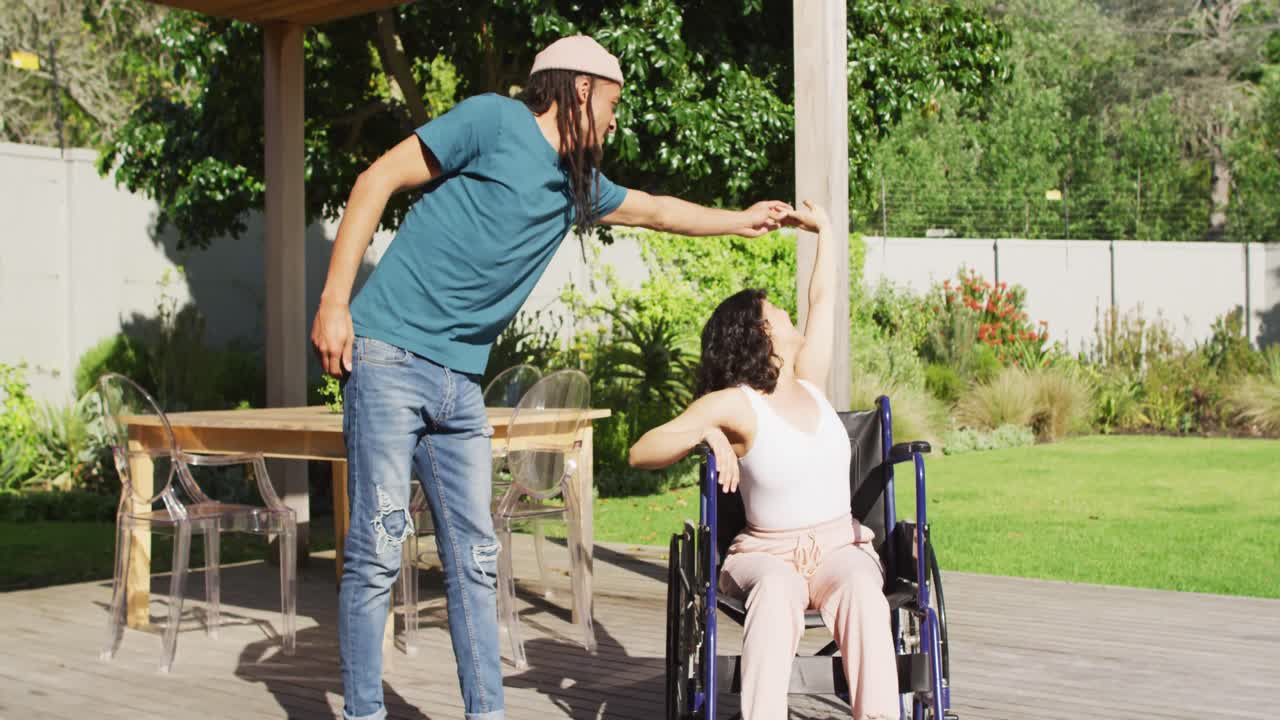 feliz pareja biracial bailando en la terraza en el jardín, mujer sentada en silla de ruedas, hombre con dreadlocks