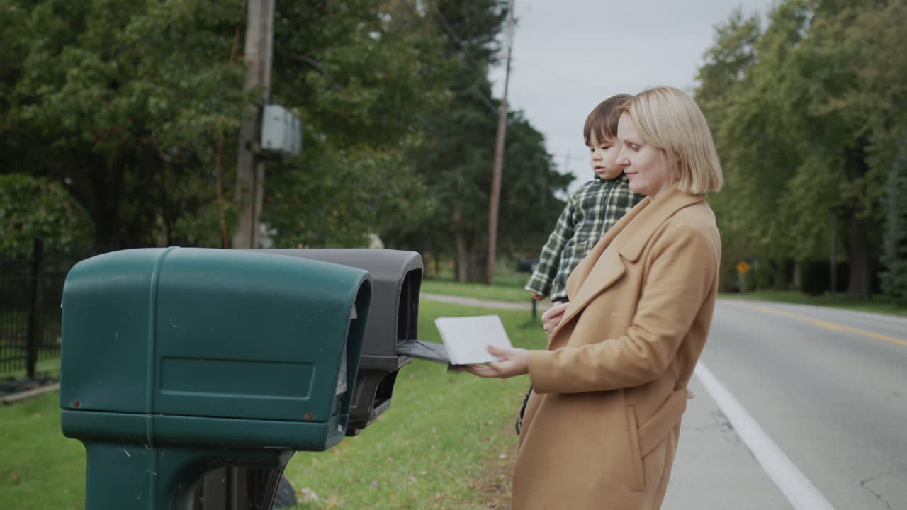 아들을 품에 들고 있는 한 여성이 편지를 보내기 위해 우편함으로 간다.