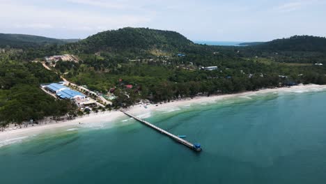 long jetty runs far into the clear blue sea from the beautiful white sand beaches with the green mountains of koh rong sanloem island in the background