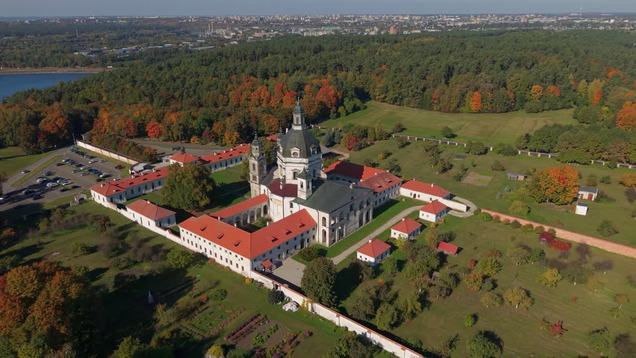 Aerial view of Pažaislis Monastery in Kaunas, Lithuania during autumn, showcasing its Baroque architecture surrounded by colorful forest and open green landscapes, zooming out