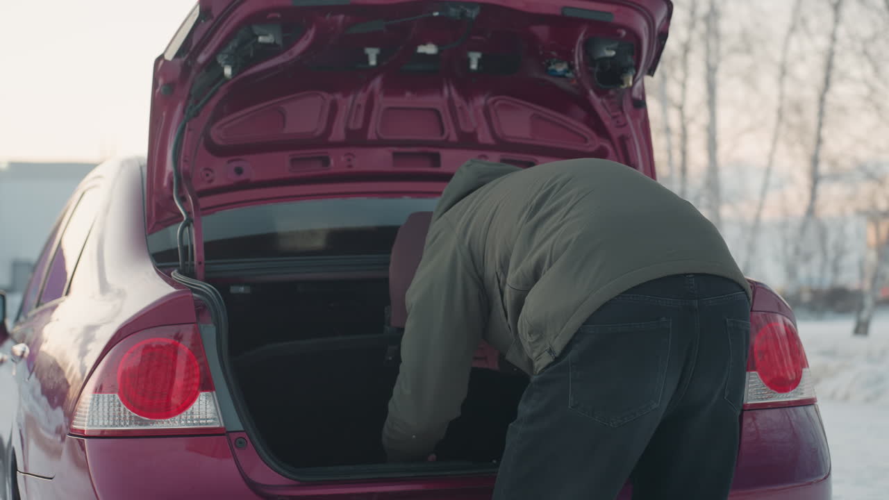 back view of student in winter outfit rummaging in car boot while parked near leafless trees and distant residential building during cold day with muted light and suburban scene