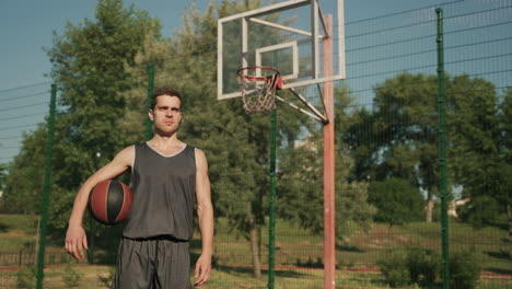 un apuesto jugador de baloncesto concentrado parado y sosteniendo la pelota en una cancha de baloncesto al aire libre en un día soleado, mirando hacia adelante con expresión enfocada y determinada