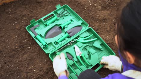 Una-Jardinera-Abre-Una-Caja-De-Herramientas-Verde-Con-Un-Juego-De-Herramientas-De-Jardinería-En-El-Suelo