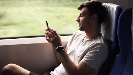 Side-view-of-a-young,-handsome-man-travels-by-modern-train.-Sitting-next-to-the-window-and-looking-at-his-mobile-phone.-Slight-natural-train-shaking.-Casual-clothes.-Nature-landscape-in-window