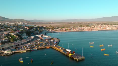 Vista-Aérea-Panorámica-Del-Muelle-Tongoy-Con-Las-Lanchas-En-El-Estacionamiento-Y-Ancladas-En-La-Orilla-Con-Las-áridas-Montañas-Del-Norte-De-Chile-Al-Fondo